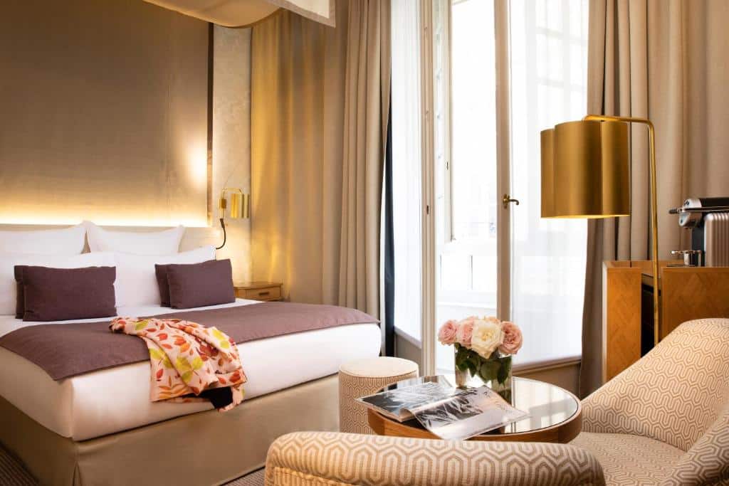 Um quarto do Hôtel D’Aubusson com uma cama de casal, poltronas, uma mesa com flores, janelas grandes e decoração neutra.