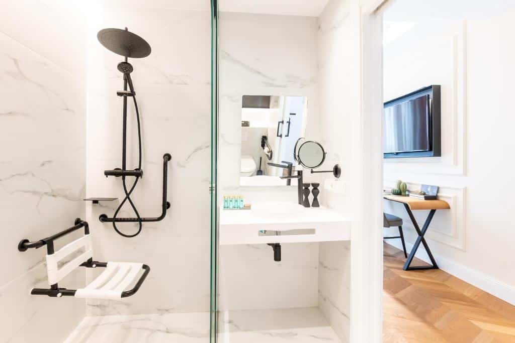 Casa de banho moderna do L&amp;H Gran Vía Valencia, com revestimento em mármore branco, chuveiro de torneira preta, espelho e prateleira com produtos de higiene.