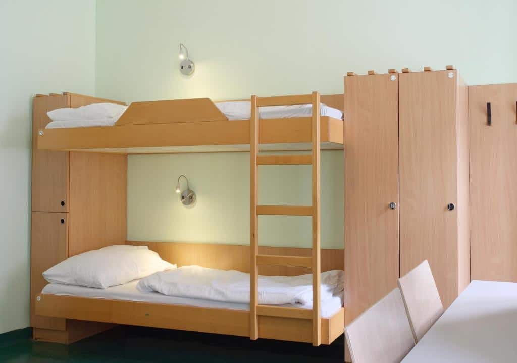 Quarto no Jugendherberge Wien – Myrthengasse, com beliches de madeira, roupa de cama branca, dois roupeiros e duas cadeiras junto a uma parede branca.