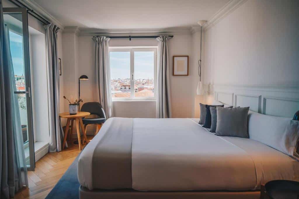 Quarto do Dear Hotel Madrid, um dos melhores hotéis em Madrid. Uma cama de casal com roupa de cama branca e cinzenta fica em frente a uma porta de vidro aberta com cortinas cinzentas dos lados. Ao fundo, uma janela de vidro com cortinas cinzentas tem vista para a cidade, e há uma mesinha com vaso de plantas, candeeiro e cadeira do lado esquerdo. Do lado direito, um quadro está pendurado na parede.