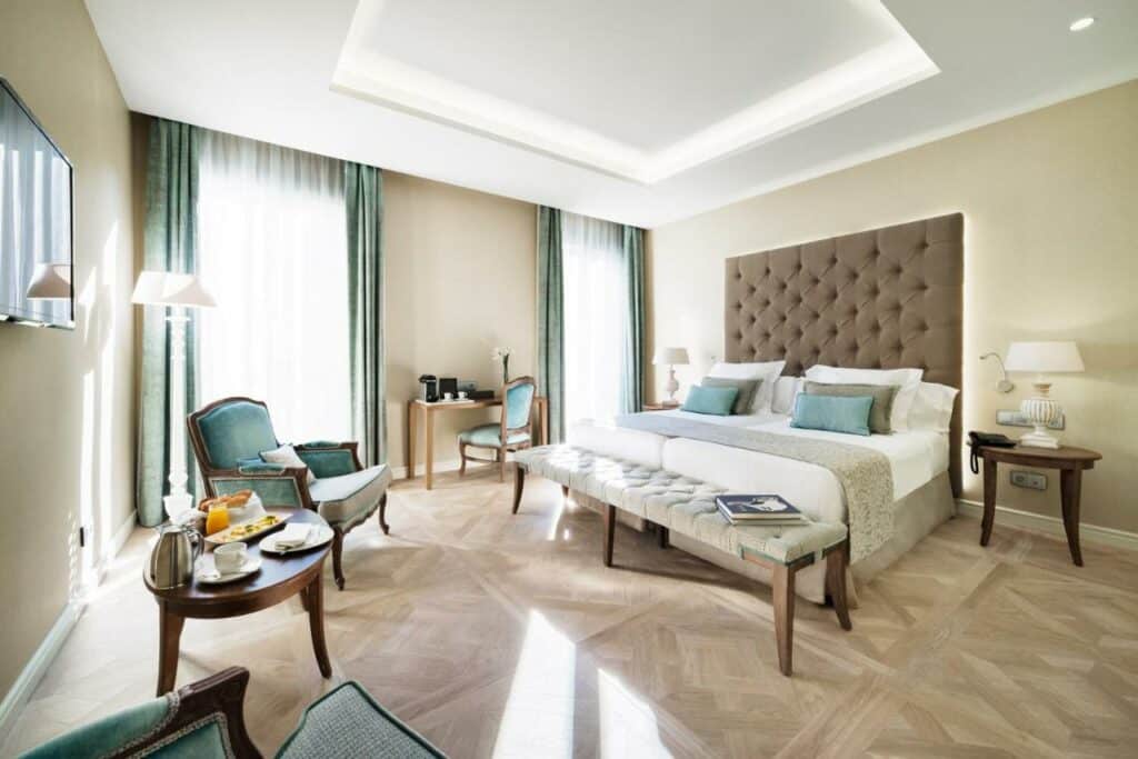 Quarto do Colón Hotel Barcelona. Uma cama de casal com candeeiros nas mesas de cabeceira, mesa de pequeno-almoço com dois cadeirões azuis, televisão e secretária junto a janelas com cortinas azuis.