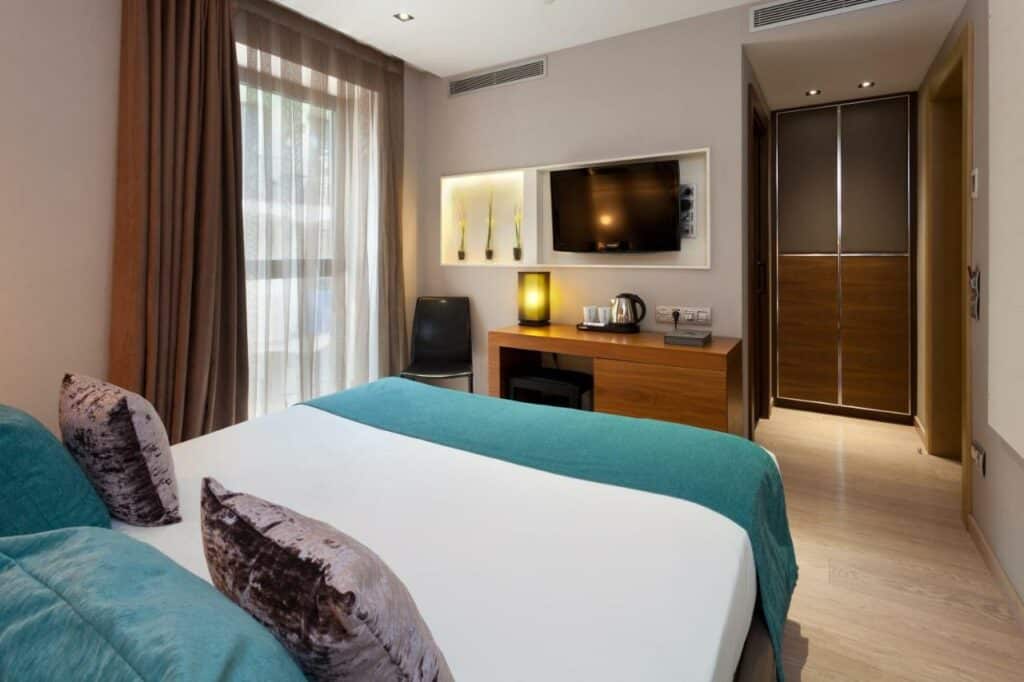 Quarto do Hotel España Ramblas, com cama de casal, televisão na parede sobre uma secretária e uma cadeira junto à janela com cortinas castanhas e brancas.