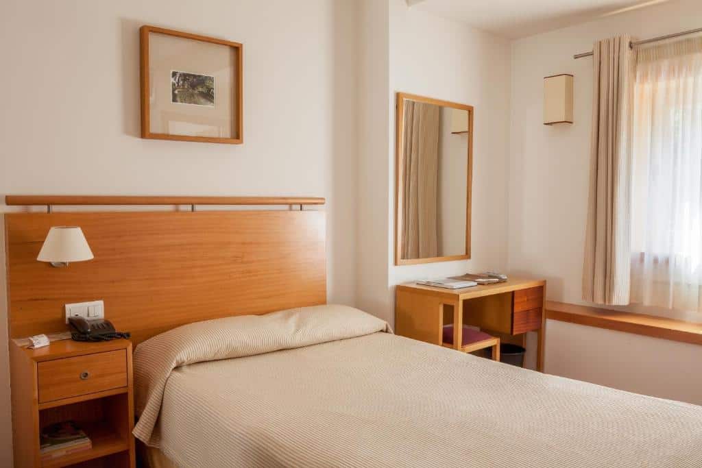 Quarto pequeno do Hotel do Lago, com cama de solteiro, cabeceira de madeira, secretária, espelho, janela com cortinas e quadro na parede.