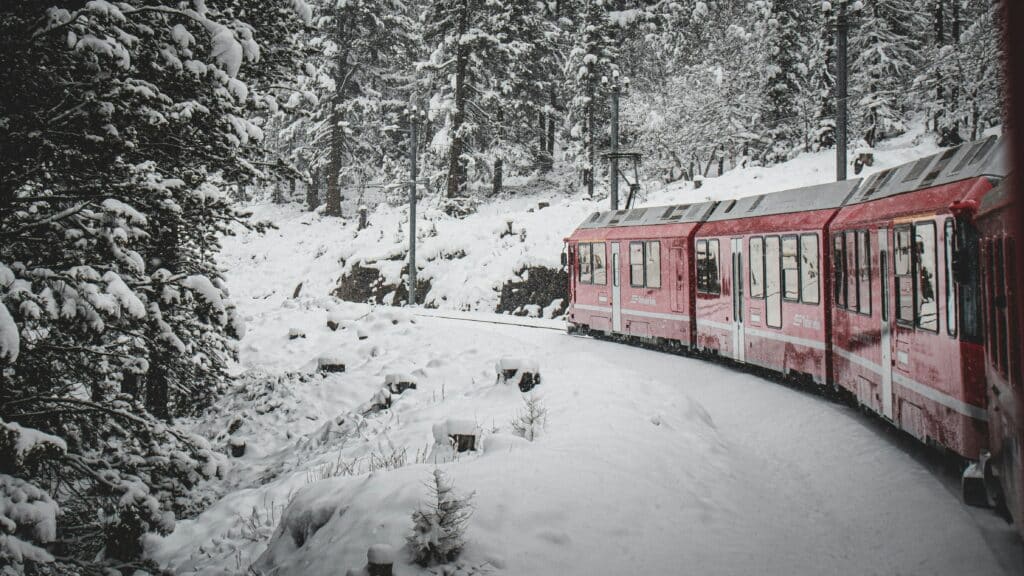 Um comboio vermelho em St. Moritz percorre uma paisagem de floresta nevada, rodeado de árvores cobertas de neve. Representa como viajar de comboio pela Suíça.