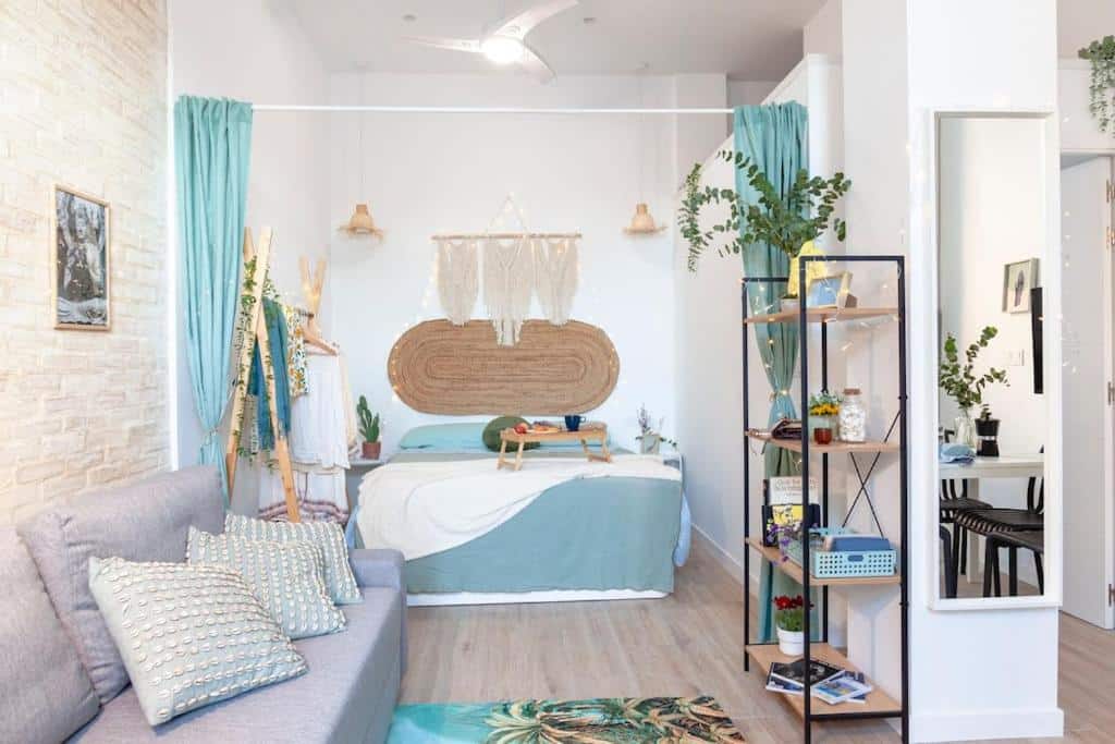 Quarto de estilo boho chic do AL5 Seaside Bliss – stylish loft, com cama de casal, cortinas, estante com plantas e vários itens de decoração em tons de azul e bege.