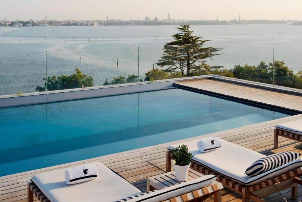 Piscina com vista panorâmica para a paisagem urbana de Veneza, com edifícios históricos e árvores ao fundo, e espreguiçadeiras de madeira com toalhas e plantas decorativas