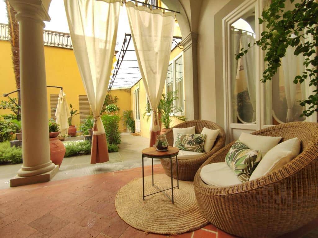 Área exterior do Renascentia Garden Relais, um dos hotéis na Toscana, com duas poltronas e três almofadas, mesa redonda pequena ao centro, cortinas e muitas flores e jardim em redor
