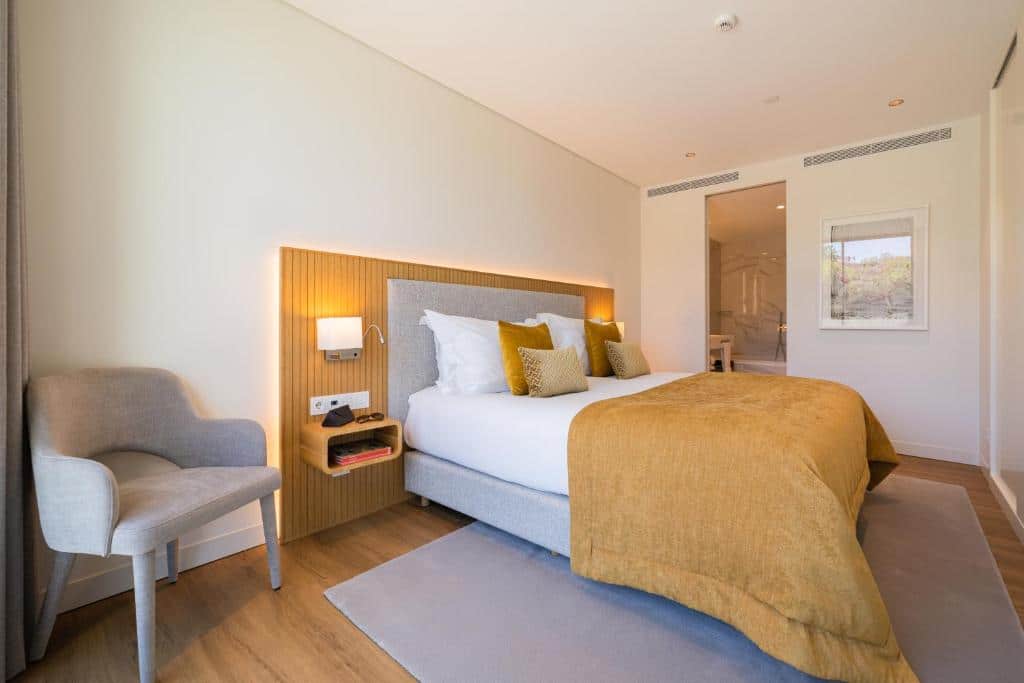 Quarto do Martinhal Lisbon Oriente com uma cama de casal ampla, uma cadeira estofada, candeeiros nas laterais da cama e uma porta que dá para uma casa de banho