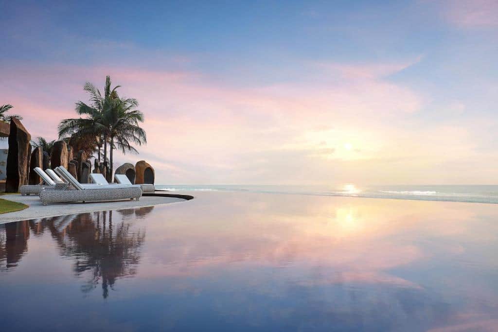 Piscina infinita ao pôr do sol, com espreguiçadeiras e palmeiras e vista para o oceano, no The Royal Purnama, em Bali.
