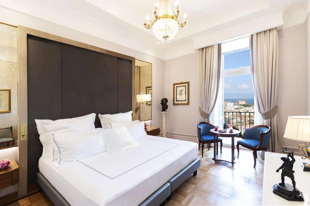 Quarto do Grand Hotel Parker's com cama de casal de lençóis brancos, duas poltronas azuis, mesa de centro e cortinas no mesmo tom, lustres e quadros num ambiente sofisticado com vista sobre a cidade.
