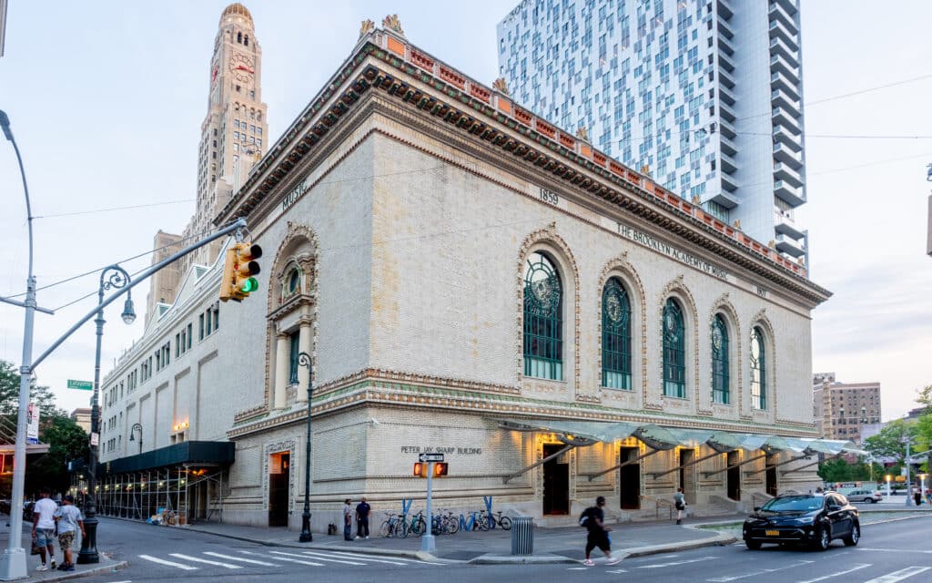 Fachada de tijolo do Brooklyn Academy of Music, com janelas em arco e pessoas a caminhar no passeio.