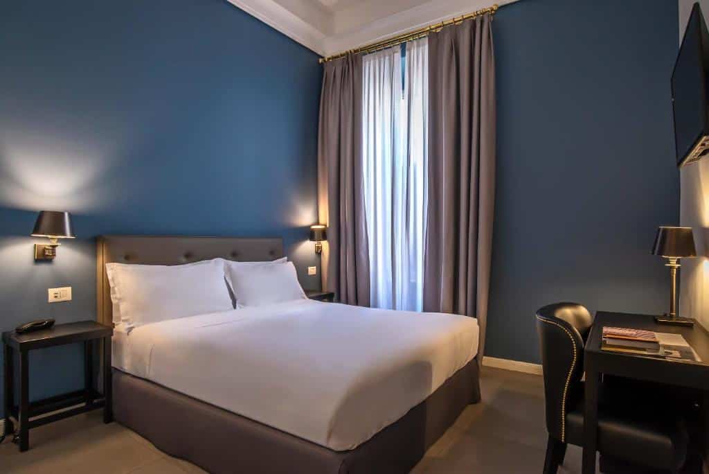 Quarto do Boutique Hotel Metro 900 com paredes azuis, cortinas cinzentas, cama de casal de roupa branca, candeeiros dourados, secretária preta e cadeira de couro.