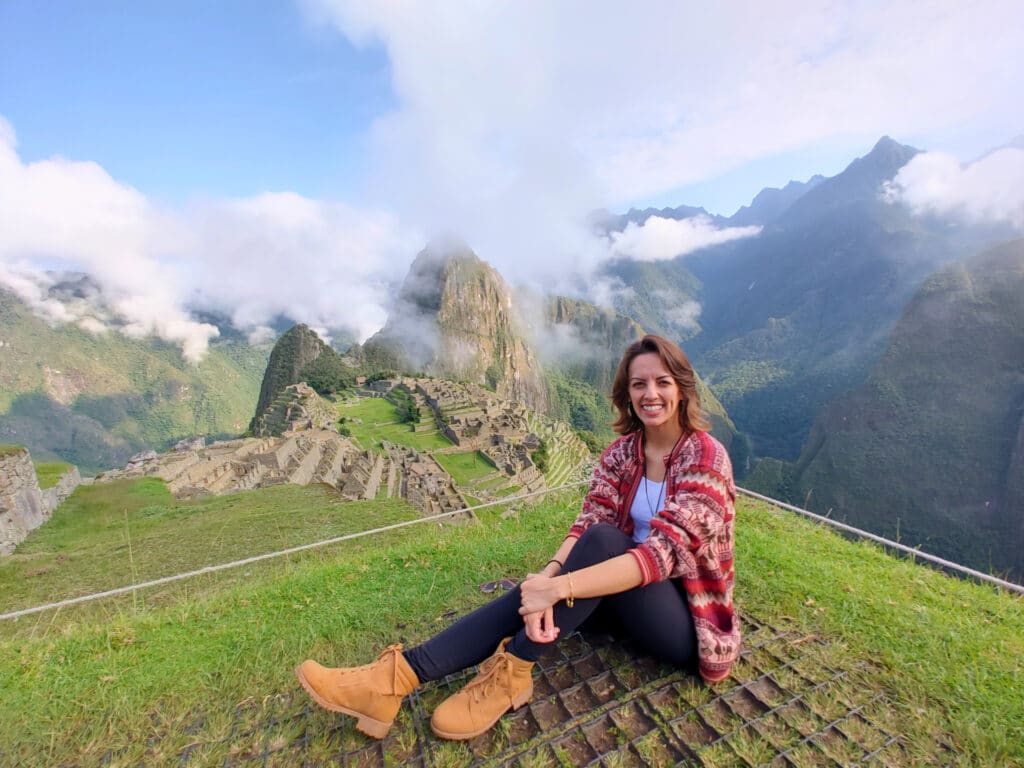 Visitante sentada na relva a sorrir, com a cidadela e as montanhas de Machu Picchu ao fundo