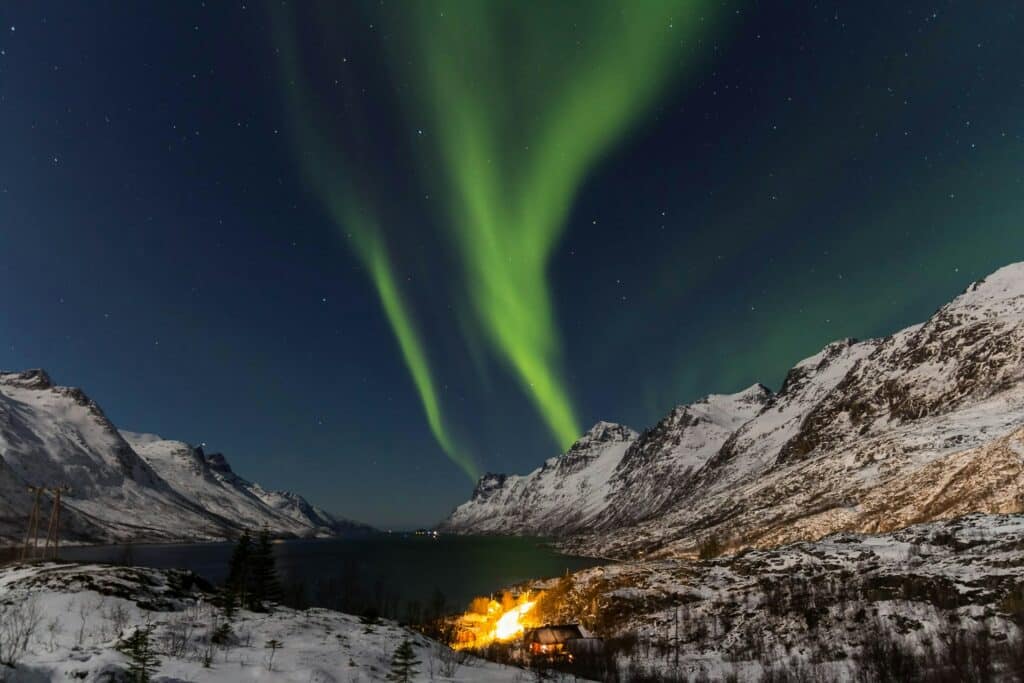 Aurora boreal a iluminar o céu noturno acima de uma paisagem montanhosa coberta de neve em Tromsø, na Noruega. Feixes de luz verde ondulante brilham intensamente contra o céu escuro estrelado, enquanto as montanhas e um lago tranquilo refletem o brilho suave.