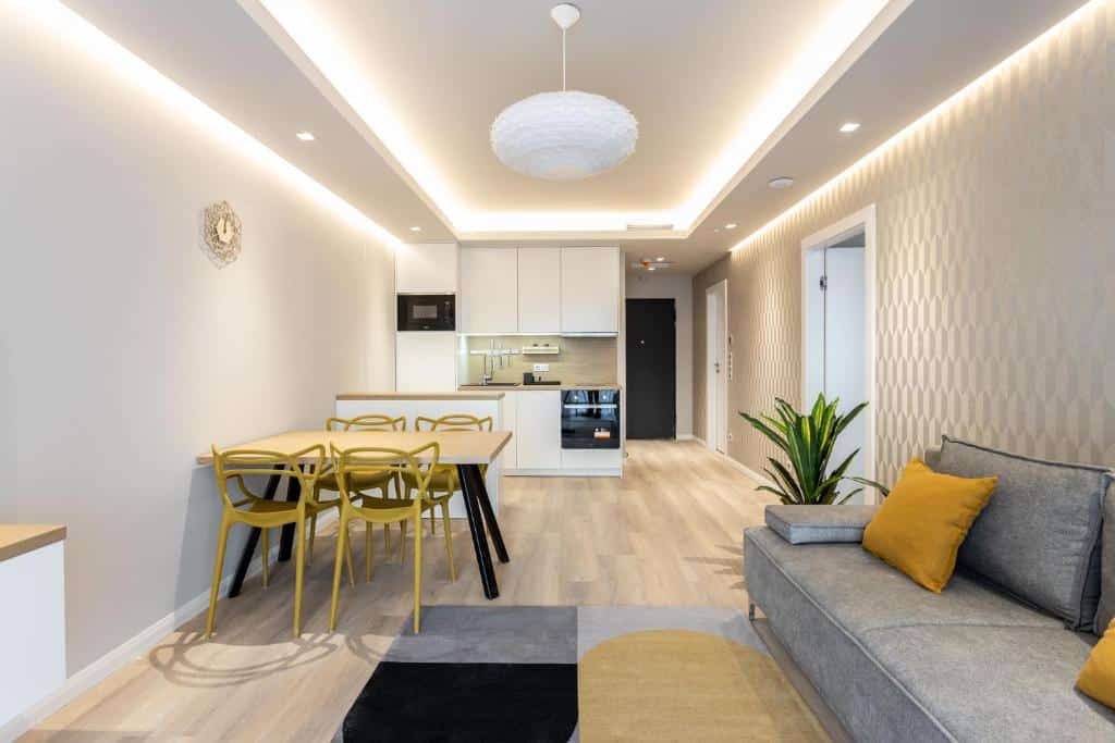 Moderna sala de estar do K46 Residence e cozinha em plano aberto, com cadeiras amarelas, um sofá cinzento e decoração neutra.