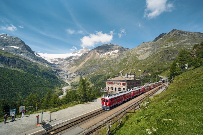 O comboio Bernina durante o dia, com a ferrovia à esquerda e montanhas ao fundo. Representa como viajar de comboio pela Suíça.