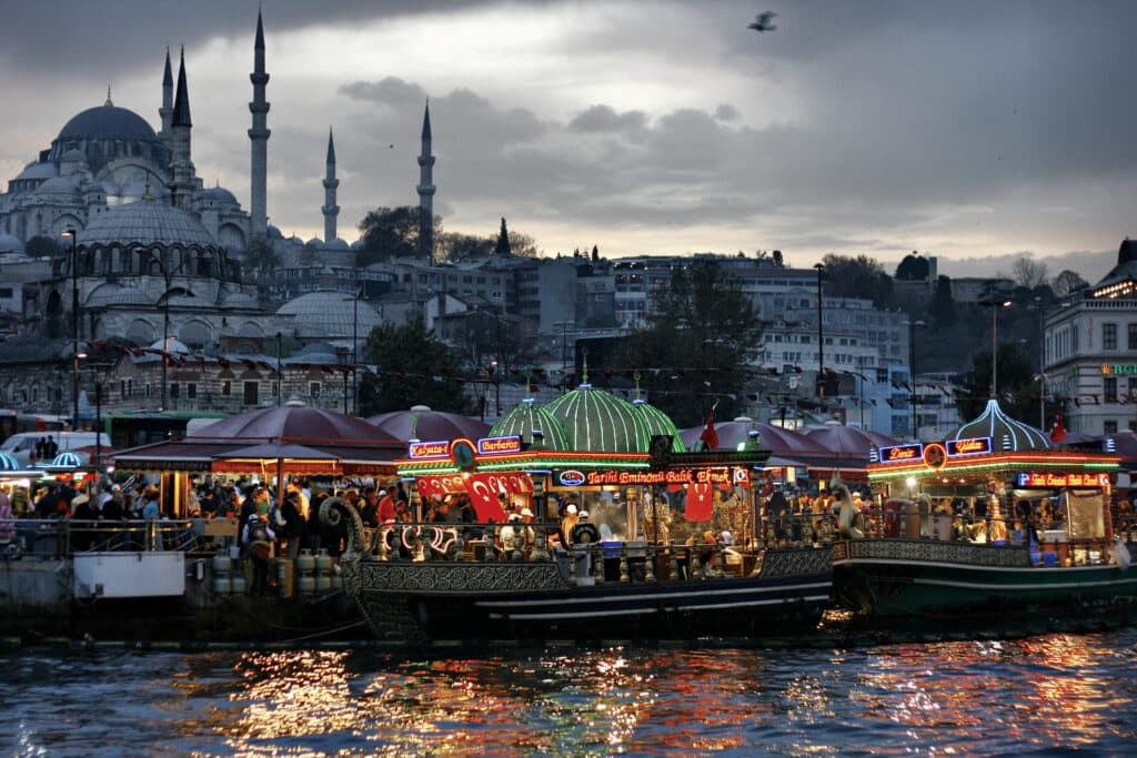 Bancas de comida coloridas e barcos alinham-se na orla de Istambul ao anoitecer, com a silhueta da mesquita ao fundo.