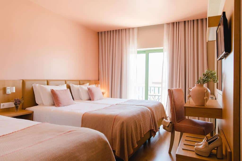 Quarto no Hotel Santo António de Fátima com duas camas de casal, paredes cor-de-rosa, roupa de cama a condizer, uma secretária com um vaso e luz do sol a entrar pela janela, para representar onde ficar em Fátima.