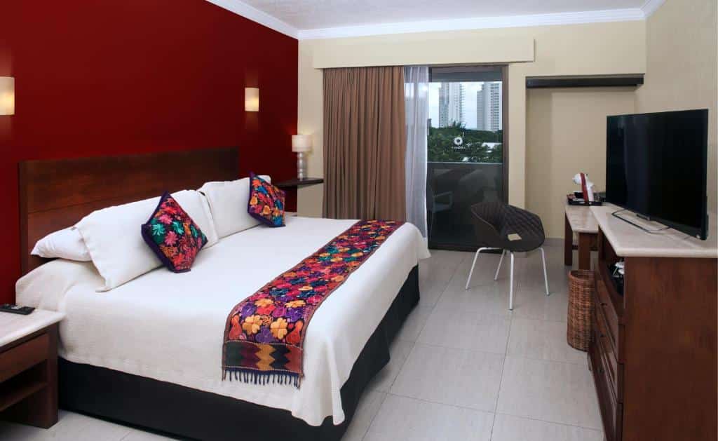 Quarto no Adhara Hacienda Cancun com cama de casal, mesa com cadeira, varanda, cómoda, televisão e duas mesas de cabeceira com candeeiros