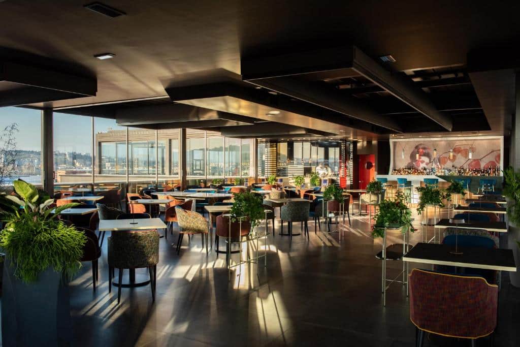 Amplo salão de restaurante com vista panorâmica sobre a cidade, decorado com mobiliário elegante e elementos naturais, num ambiente moderno e sofisticado do Gold Tower Lifestyle Hotel.