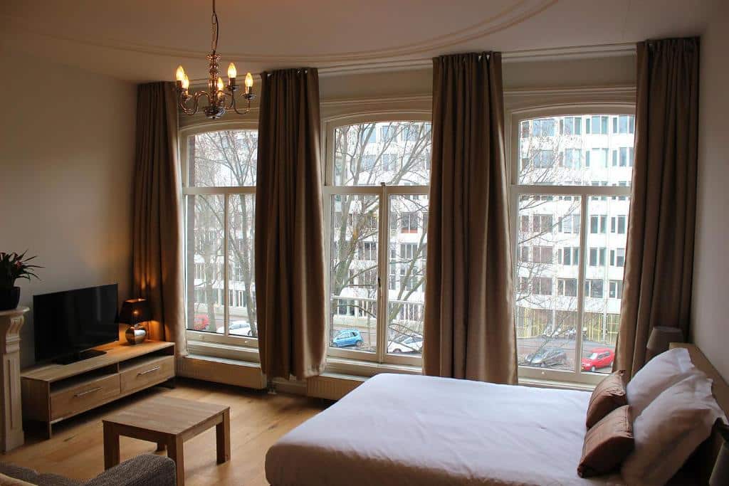 Quarto do Amsterdam Jewel Canal Apartments, que ilustra onde ficar em Amesterdão: cama grande, TV, mobiliário de madeira e três janelas altas com cortinados castanhos e vista para uma rua.