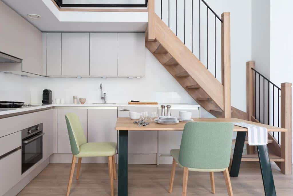 Cozinha moderna do apartamento Wittenberg by Cove, com armários de madeira clara, uma pequena mesa de refeições para quatro pessoas e uma escada de madeira com corrimão preto.