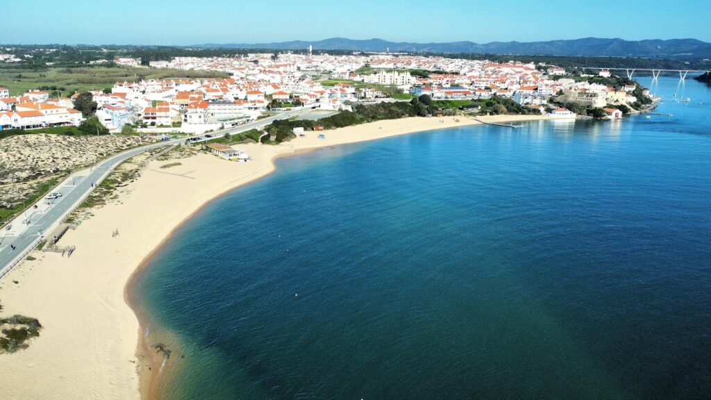 Vista aérea de uma vila costeira com edifícios brancos e telhados vermelhos, praia de areia dourada e mar azul calmo.