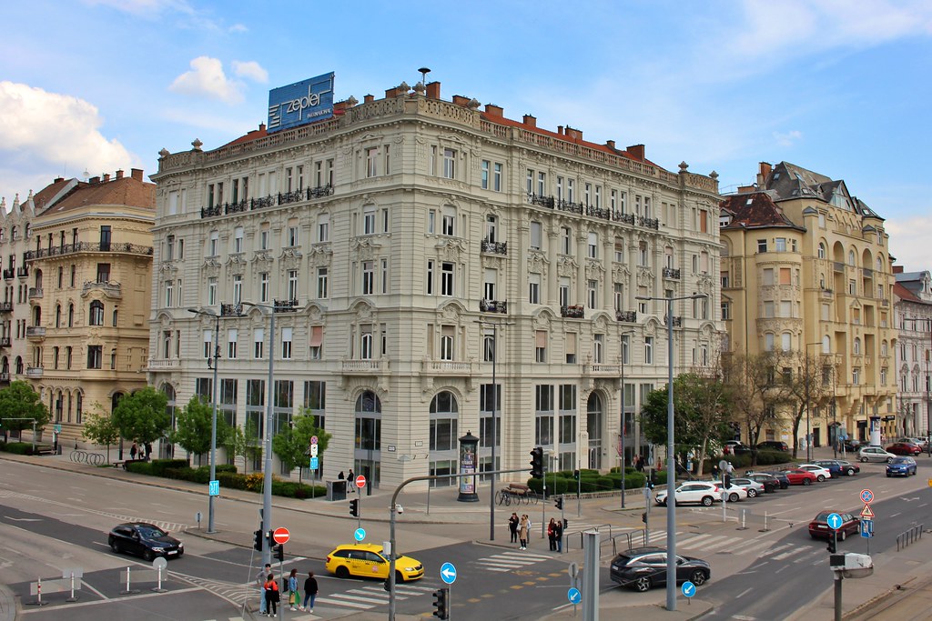 Um grande edifício em Belváros, ornamentado e de cor clara, num cruzamento movimentado da cidade, com carros, peões e placas de rua.
