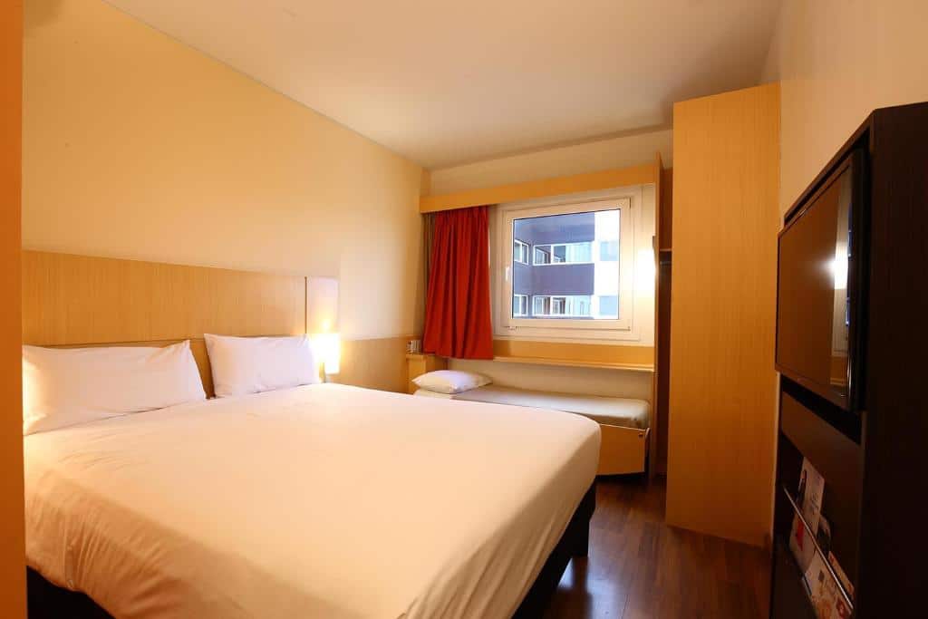 quarto compacto e funcional do ibis Buenos Aires Obelisco com cama de casal centralizada, janela com cortinas vermelhas, tv de ecrã plano na parede e um pequeno espaço de trabalho