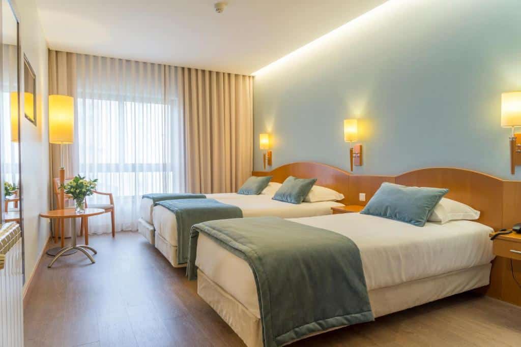 Quarto no Hotel Cinquentenário & Conference Center com duas camas de casal, apontamentos a azul, mobiliário em madeira, candeeiros de parede e uma janela com cortinados transparentes.