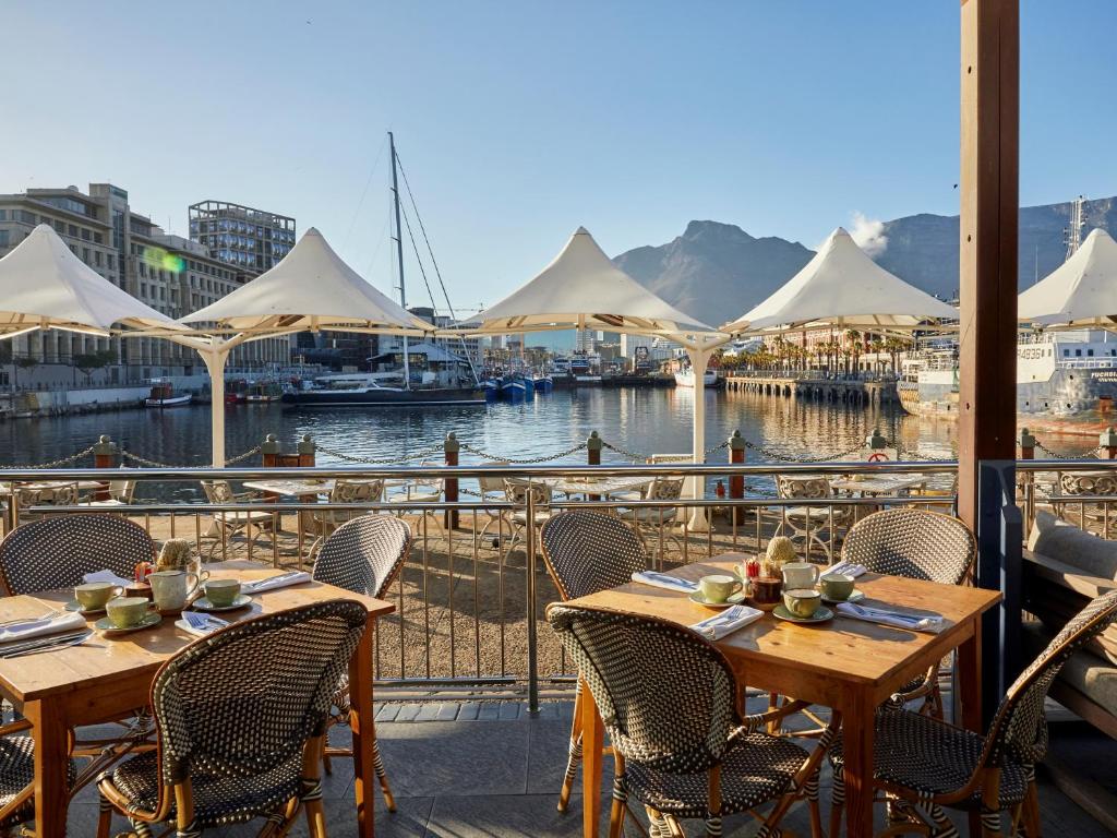 Restaurante ao ar livre no Victoria & Alfred Hotel com mesas de madeira e vista para uma marina, barcos e montanhas.