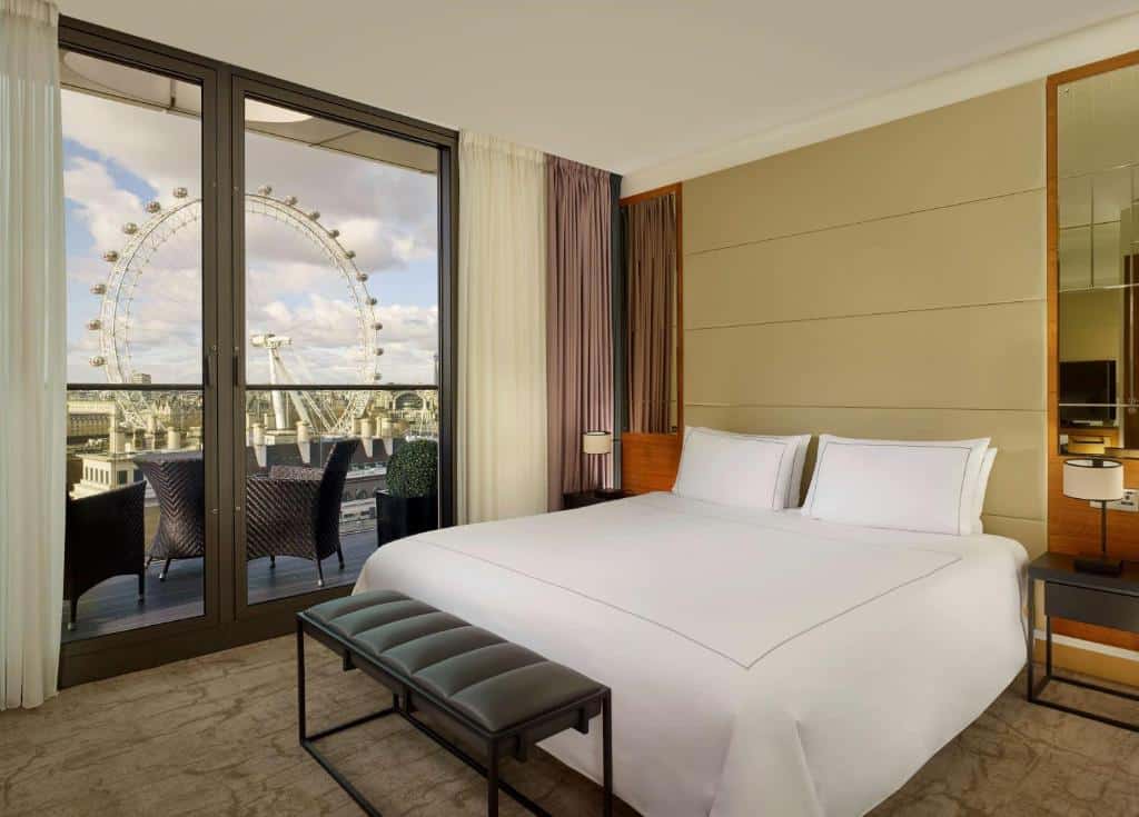 Quarto do Park Plaza County Hall London com varanda com mesa e cadeiras e vista para a London Eye, cama de casal, duas mesas de cabeceira e um pequeno banco.