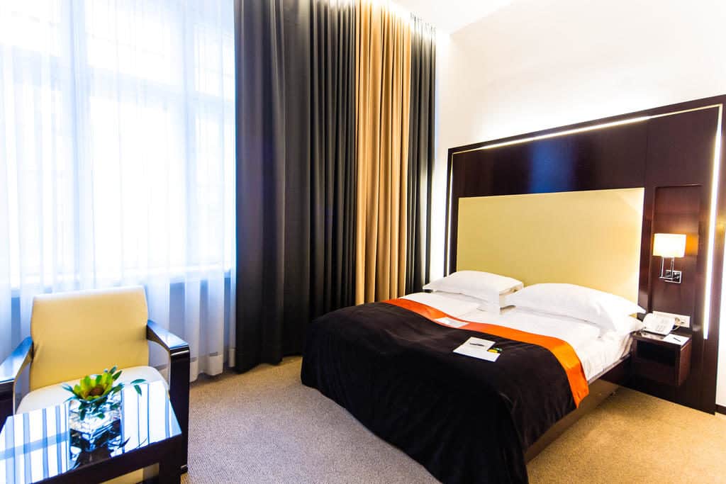 Quarto no Levante Parliament, com cama de casal, poltrona, mesa de vidro e uma janela grande com cortinados transparentes e blackout.