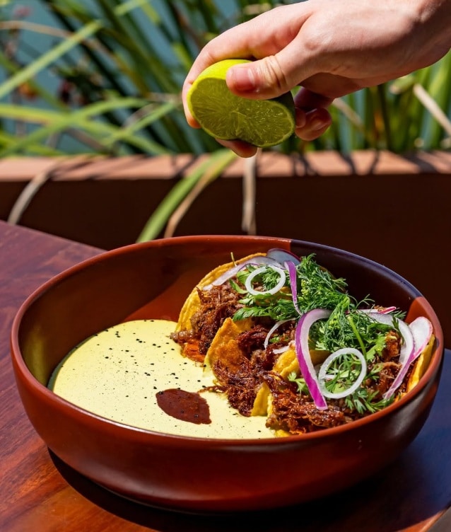 Prato de comida típica mexicana, com molho amarelo, pequenos tacos com carne desfiada, cebolinho e cebola roxa, servido em tigela de cerâmica rústica, com uma mão a segurar uma lima verde.
