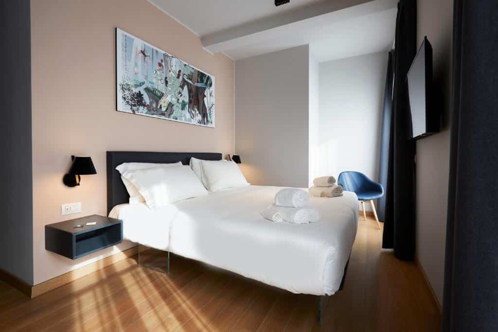 Quarto do 21 House of Stories Città Studi com cama de casal, televisão, pavimento que imita madeira, cadeira junto à janela e mesinha de cabeceira com candeeiro