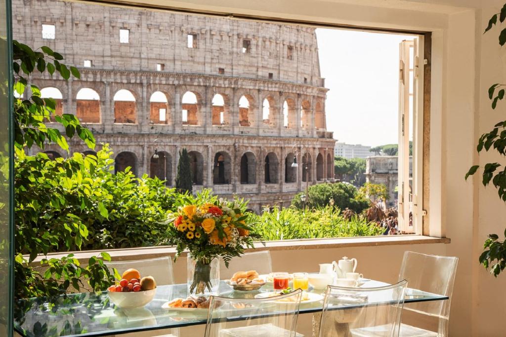 vista da sala do 47Luxury Suites – Colosseo, com o pequeno-almoço posto numa mesa de vidro, cadeiras e um vaso de flores, com vista para o Coliseu e plantas em redor