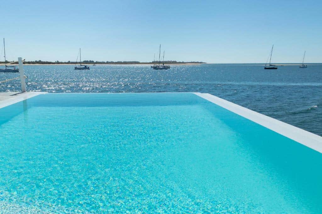 Piscina infinita com vista para um mar calmo com vários veleiros ancorados ao longe, sob um céu limpo, no Grand House – Relais & Chateaux.