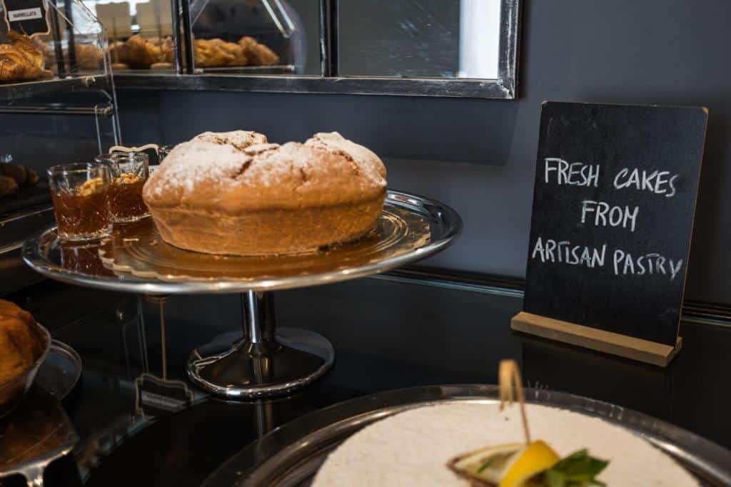 Bolo artesanal dourado polvilhado com açúcar num prato sobre o balcão de uma cafetaria, junto a uma placa de padaria artesanal, no Boutique Hotel Metro 900.