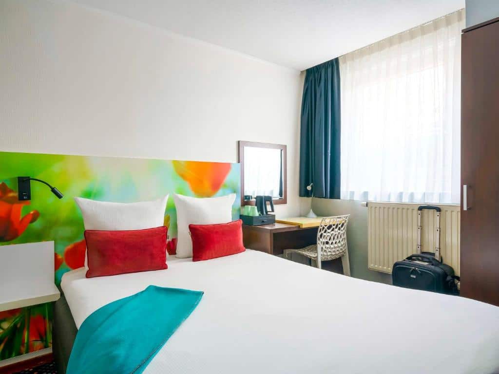 Quarto do ibis Styles Amsterdam City, que ilustra onde ficar em Amesterdão: cama de casal, cabeceira colorida, secretária, cadeira, janela com cortinados e uma mala ao lado do roupeiro.