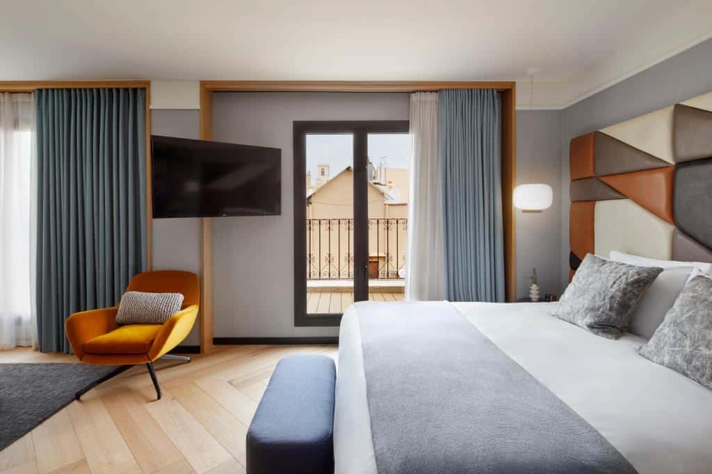 Quarto do Kimpton Vividora Hotel. A cama de casal encosta a um painel colorido, com banco aos pés, candeeiros nas mesas de cabeceira, televisão em frente e um cadeirão amarelo.