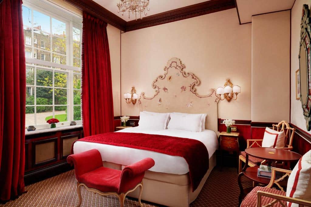 Quarto do The Montague On The Gardens com cama de casal e cabeceira clássica, uma janela ampla com cortinas vermelhas e uma mesa redonda com duas cadeiras, decorado em vermelho, branco e dourado.