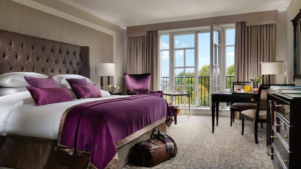 Quarto de hotel elegante com cama king size, cabeceira estofada, cortinas e pormenores em tons de roxo, janelas amplas com vista para o exterior e secretária com portátil.