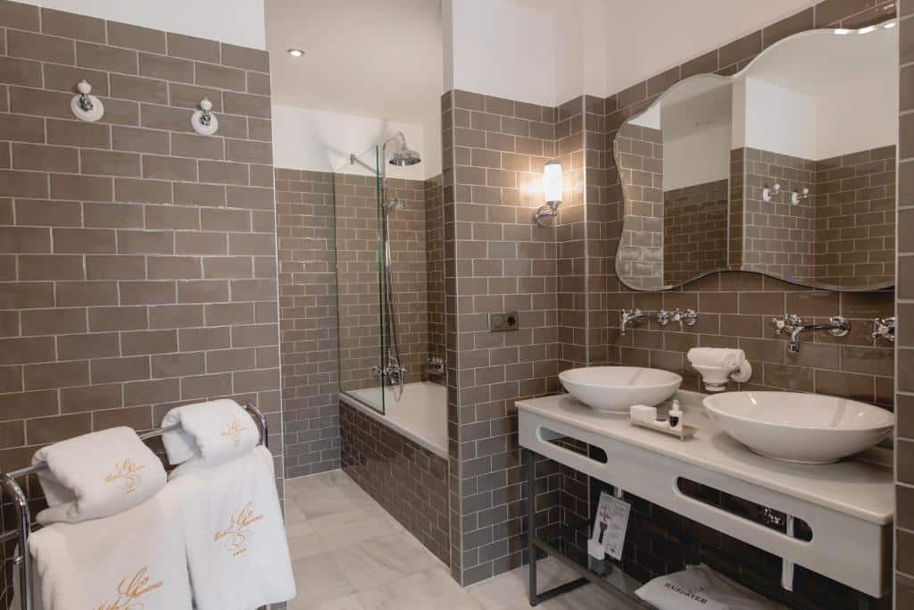 Casa de banho do Hotel Gravina 51 com dois lavatórios e espelho, toalhas à esquerda e banheira com chuveiro ao fundo.