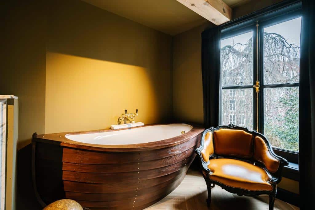 Banheira do Quarto Duplo Grand Deluxe do Boutique Hotel The Noblemen, revestida a madeira, com uma cadeira de época ao lado e janela com vista para árvores