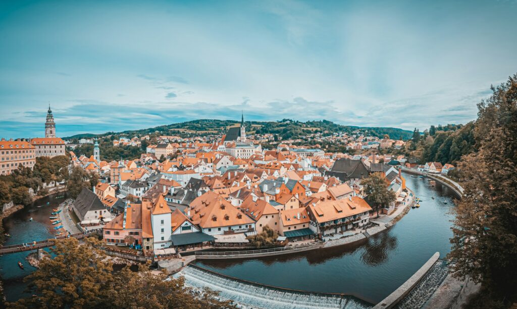 Vista aérea panorâmica de Český Krumlov, na República Checa, mostrando a cidade histórica com telhados cor de laranja em torno de um meandro do rio Vltava. Há vegetação verde e colinas ao fundo. No centro da imagem, destaca-se uma torre de igreja pontiaguda.