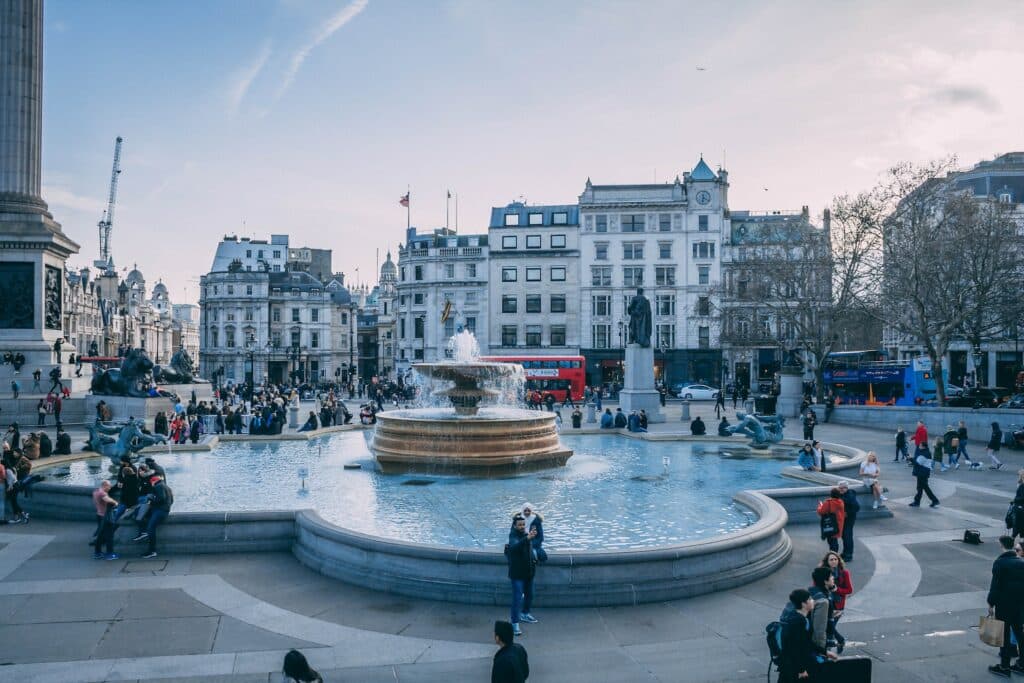 Praça pública na Trafalgar Square com fontes arredondadas e um repuxo, algumas pessoas a passear e edifícios antigos em redor
