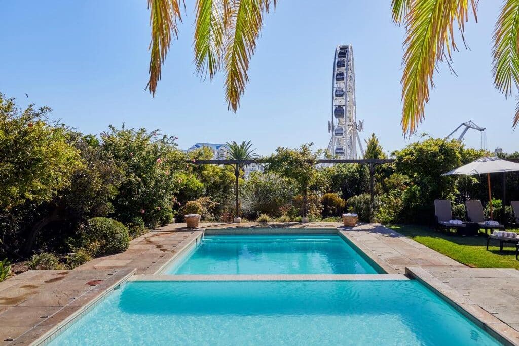 Piscina no Queen Victoria Hotel com espreguiçadeiras, vegetação e uma roda-gigante ao fundo sob o céu azul.