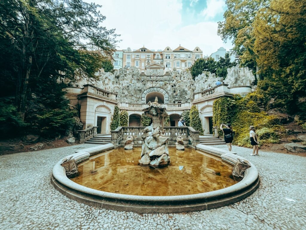 Uma fonte de pedra com estátuas num tanque redondo em frente a um grande edifício e a um jardim rochoso, com pessoas a tirar fotografias.
