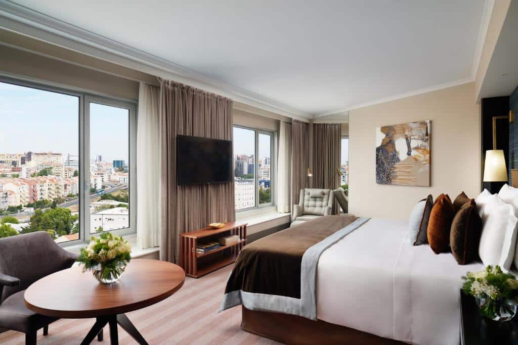 Quarto do Corinthia Lisbon com duas janelas amplas com vista para a cidade em frente à cama de casal, uma televisão entre as janelas, uma poltrona e uma pequena mesa com cadeira estofada