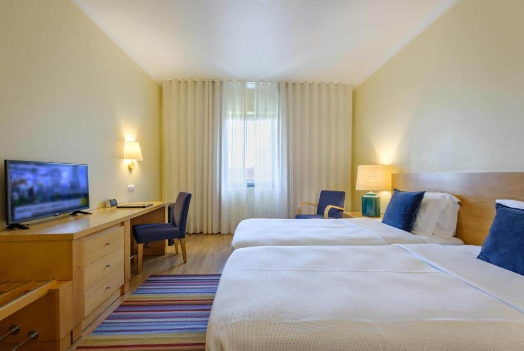 Quarto do Hotel Santa Maria com duas camas de solteiro, uma secretária com cadeira, TV, poltrona, candeeiro e janela com cortinas brancas, para representar onde ficar em Fátima.